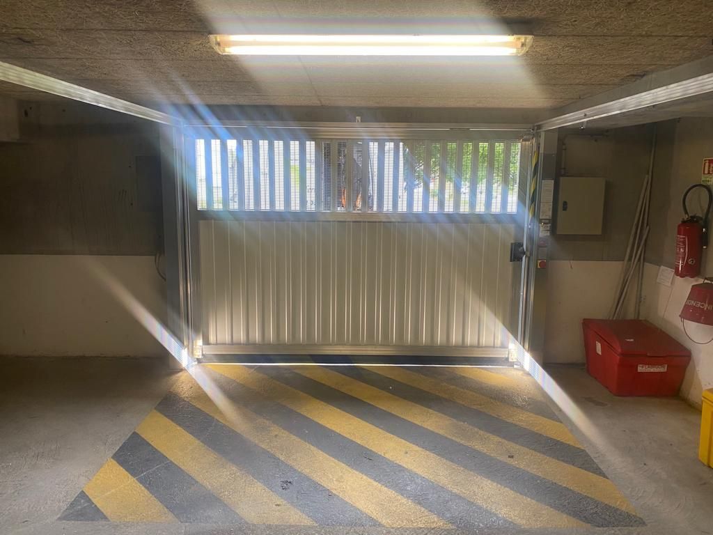 Entrée d'un parking souterrain avec portail blanc, sol à rayures jaunes et éclairage fluorescent au plafond