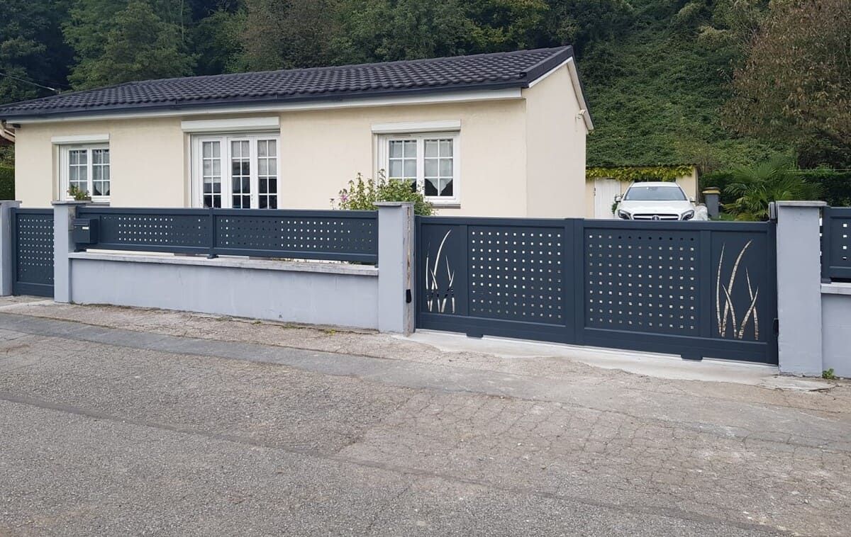 Maison beige de plain-pied avec une clôture et un portail gris foncé, donnant sur une rue de gravier.