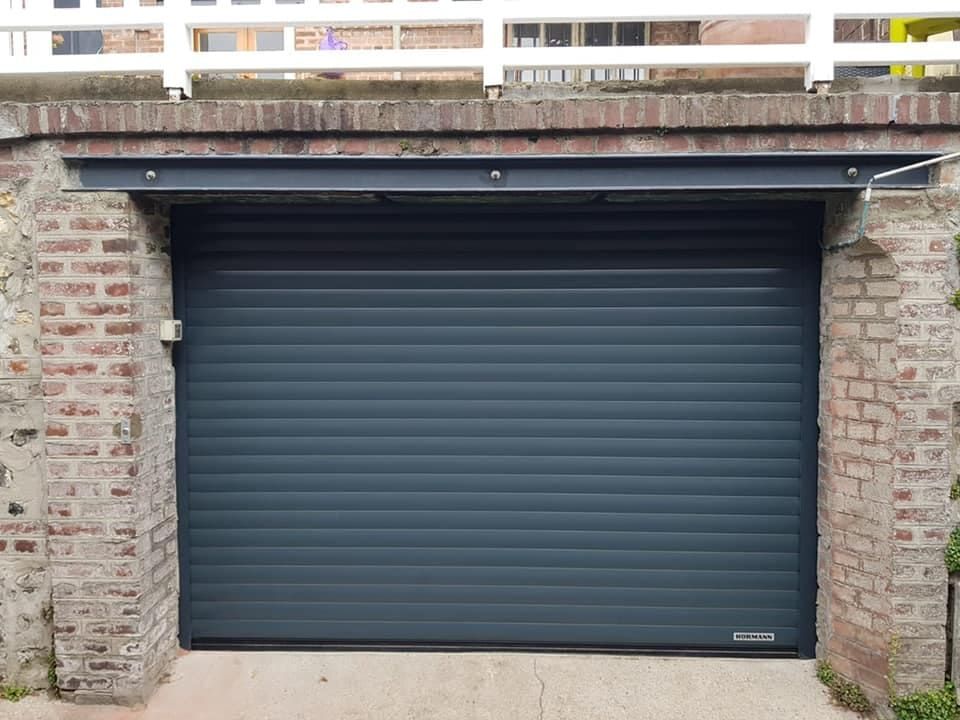 Porte de garage fermée, gris foncé, encastrée dans un mur de briques sous un auvent en bois.
