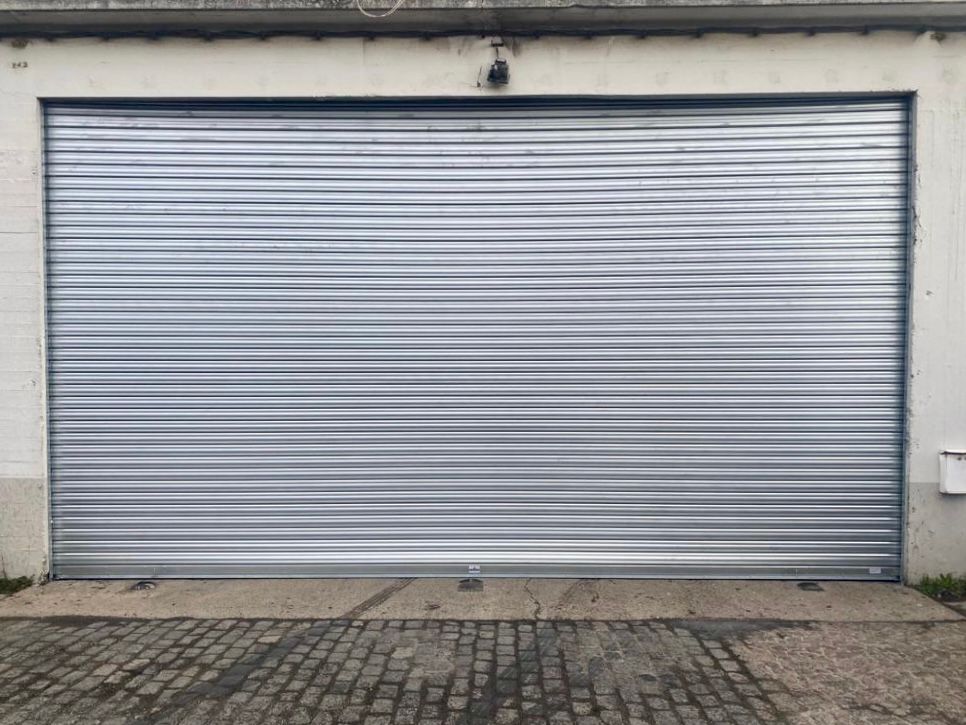 Porte de garage métallique argentée fermée sur un mur blanc, avec une allée pavée devant.