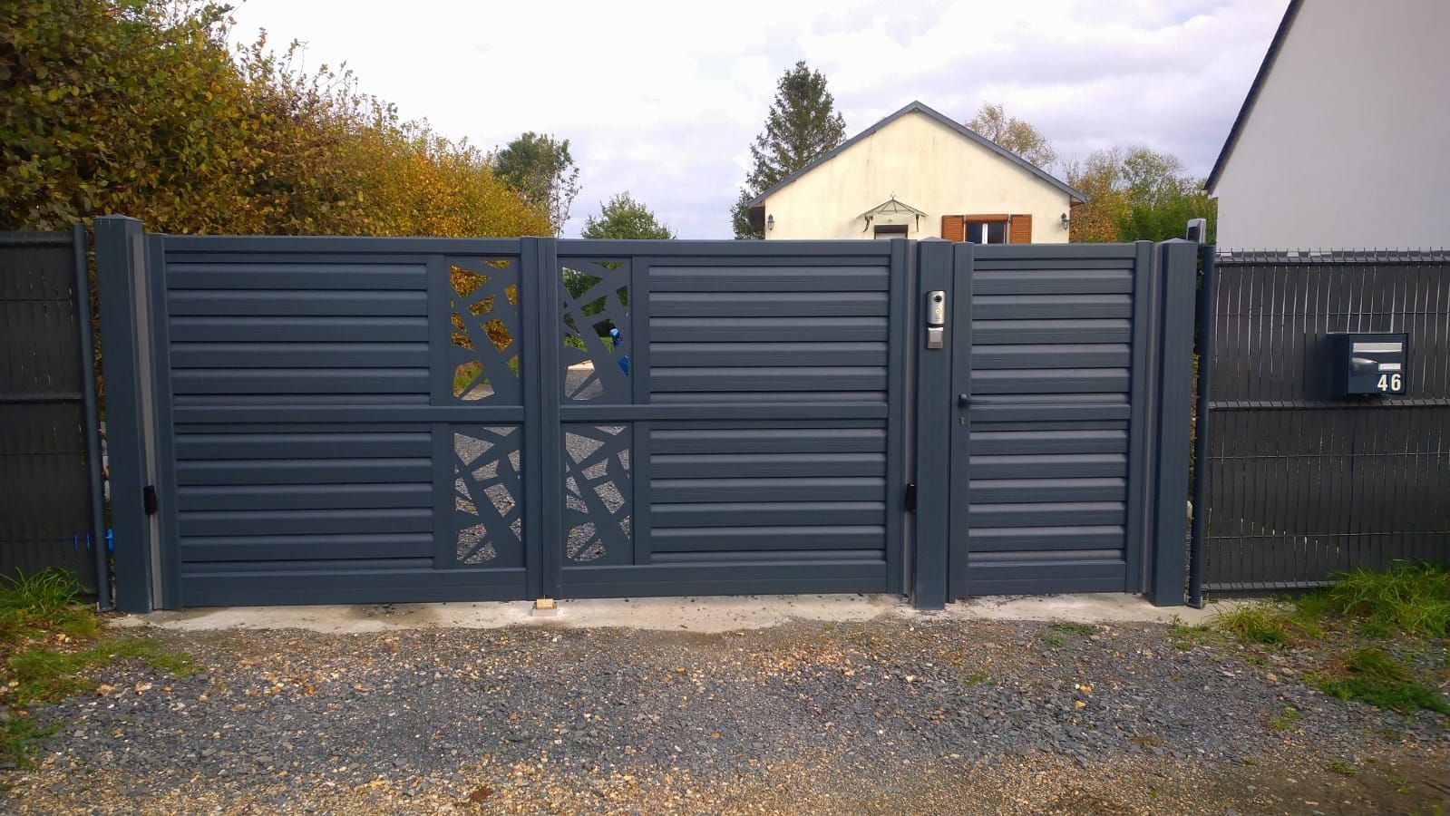 Portail d'allée en métal gris avec découpes décoratives devant une maison et une route de gravier