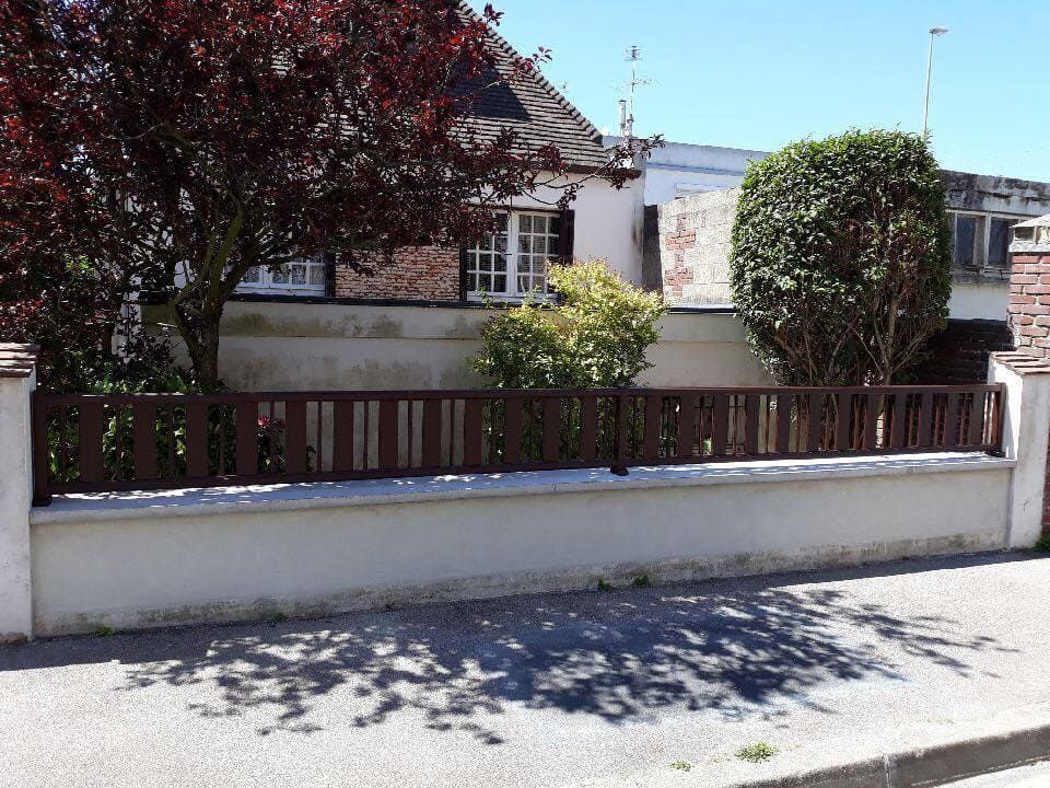 Maison résidentielle tranquille derrière un muret blanc et une clôture en bois, les arbres projetant des ombres sur la rue.