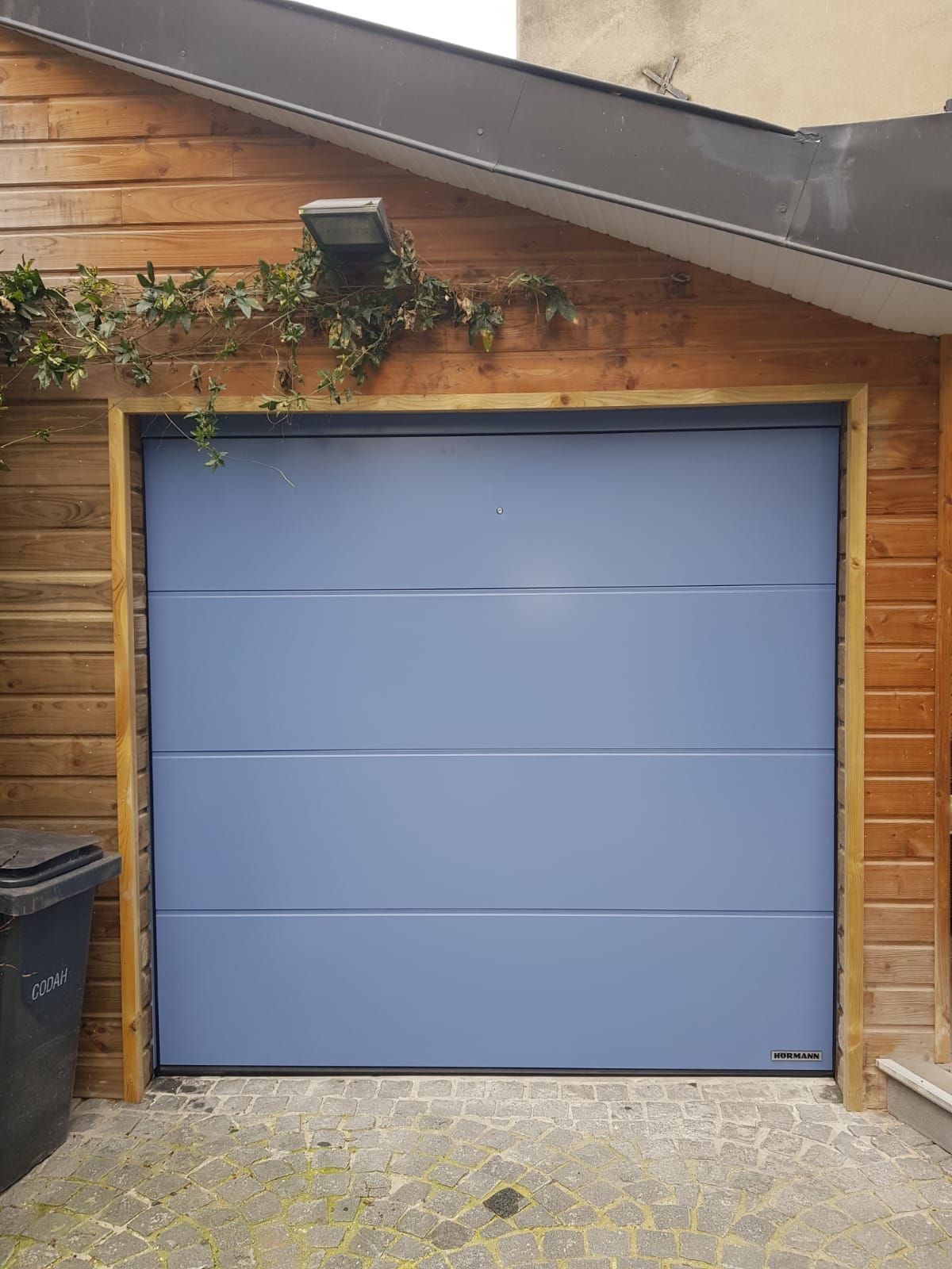 Porte de garage bleue sur la façade d'une maison en bois, avec du lierre au-dessus et une poubelle sur la gauche.
