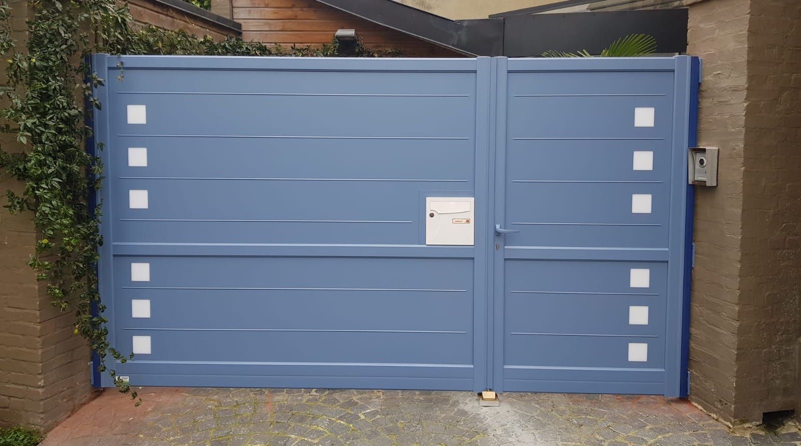 Porte de garage double bleue avec petites fenêtres carrées et boîte aux lettres, encastrée dans un mur de briques.