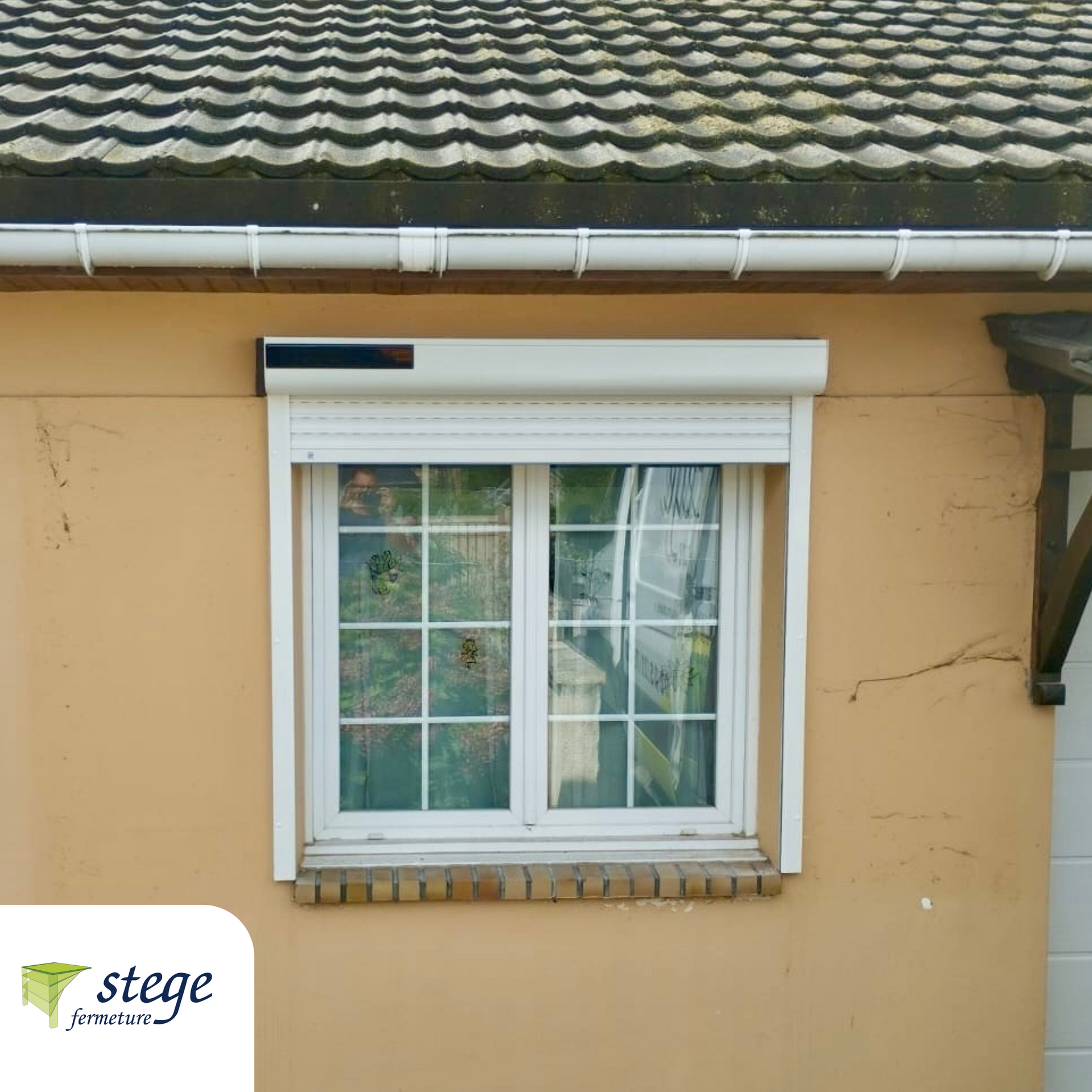 Fenêtre à cadre blanc avec volet roulant sur un mur beige d'une maison sous un toit de tuiles.