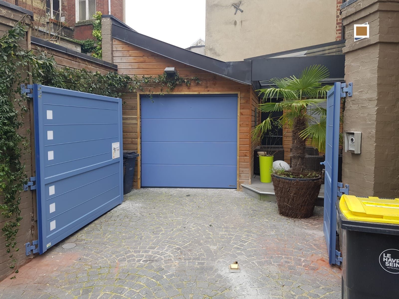 Des portes de garage bleues dans une allée de cour, bordée de murs en briques et de plantes en pot.