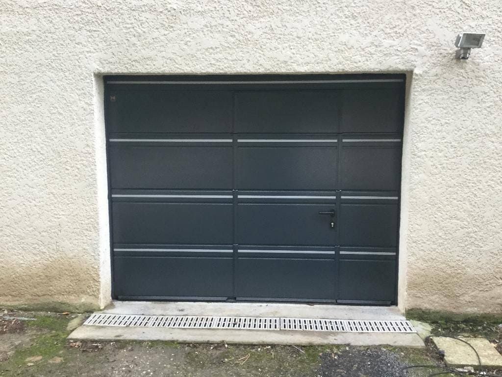 Porte de garage noire encastrée dans un mur en stuc blanc, au-dessus d'un caniveau en béton.