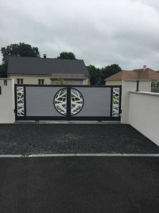 Portail d'allée gris à motif circulaire devant une maison sous un ciel nuageux