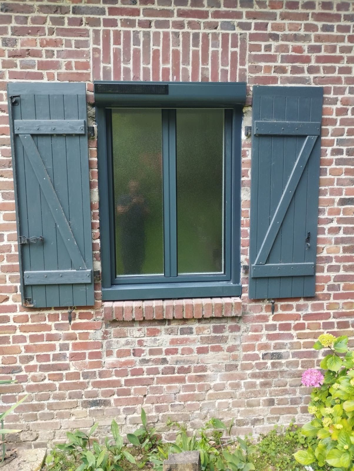 Fenêtre d'une maison en briques avec volets vert foncé et parterre de fleurs en dessous