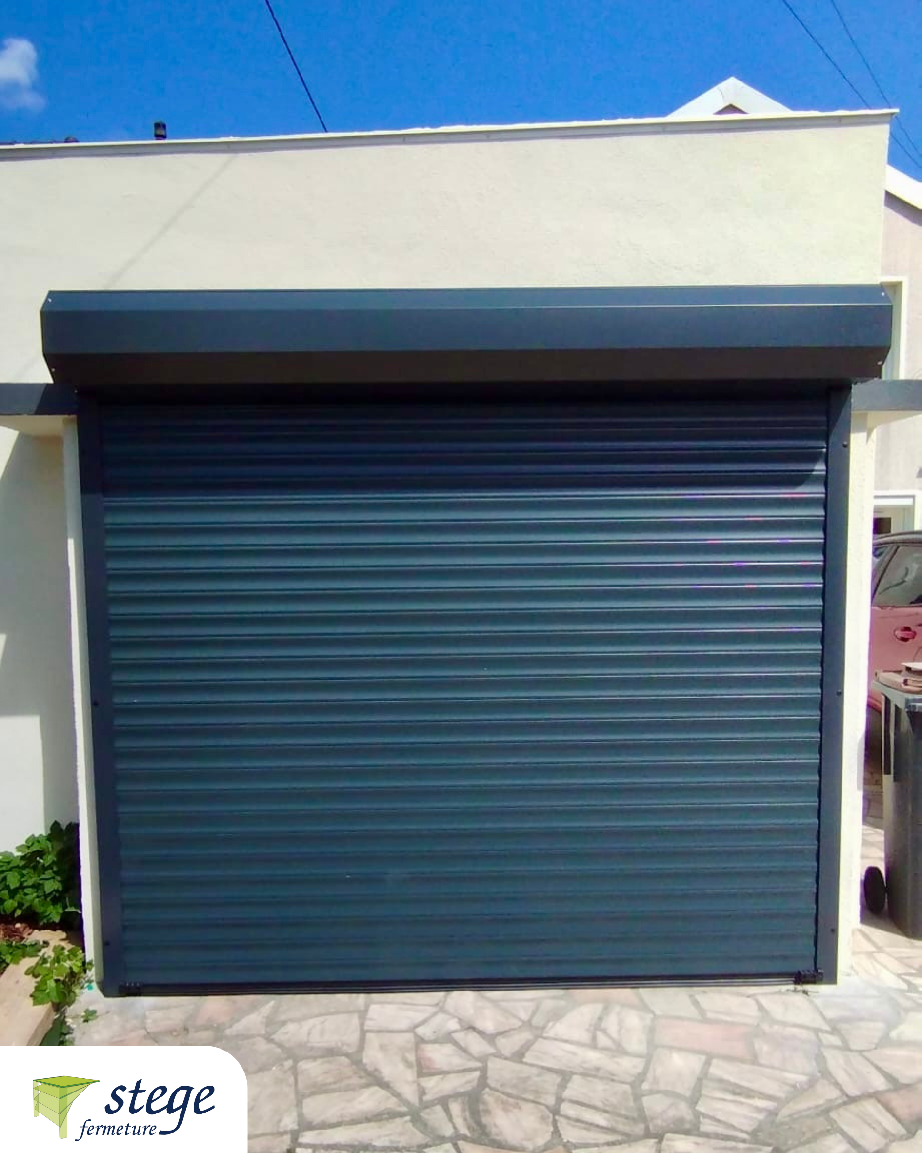 Porte de garage sectionnelle en métal bleu sur un bâtiment beige avec une terrasse en pierre devant