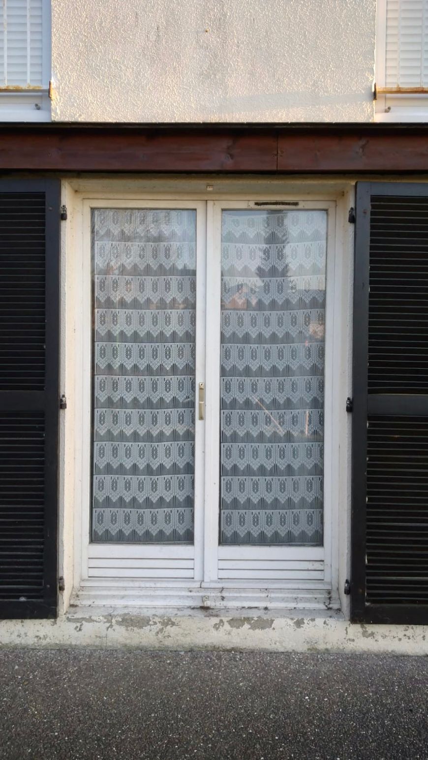 Double porte-fenêtre blanche avec rideaux de dentelle entre des volets noirs sur une façade d'immeuble beige