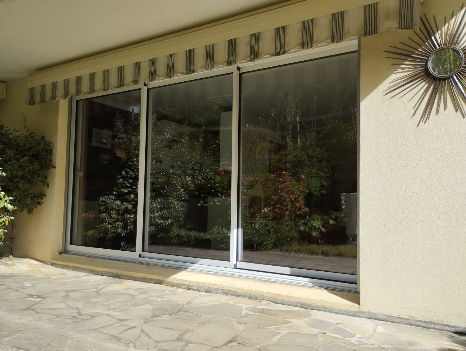 Portes coulissantes en verre donnant sur une terrasse avec store rayé et pavage en pierre, reflétant la verdure extérieure