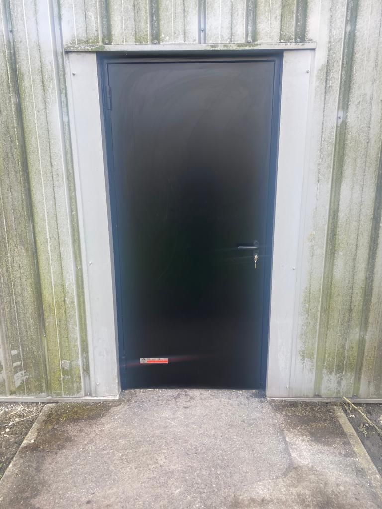 Porte en métal noir dans un mur en béton patiné, avec un petit panneau rouge près du bas.