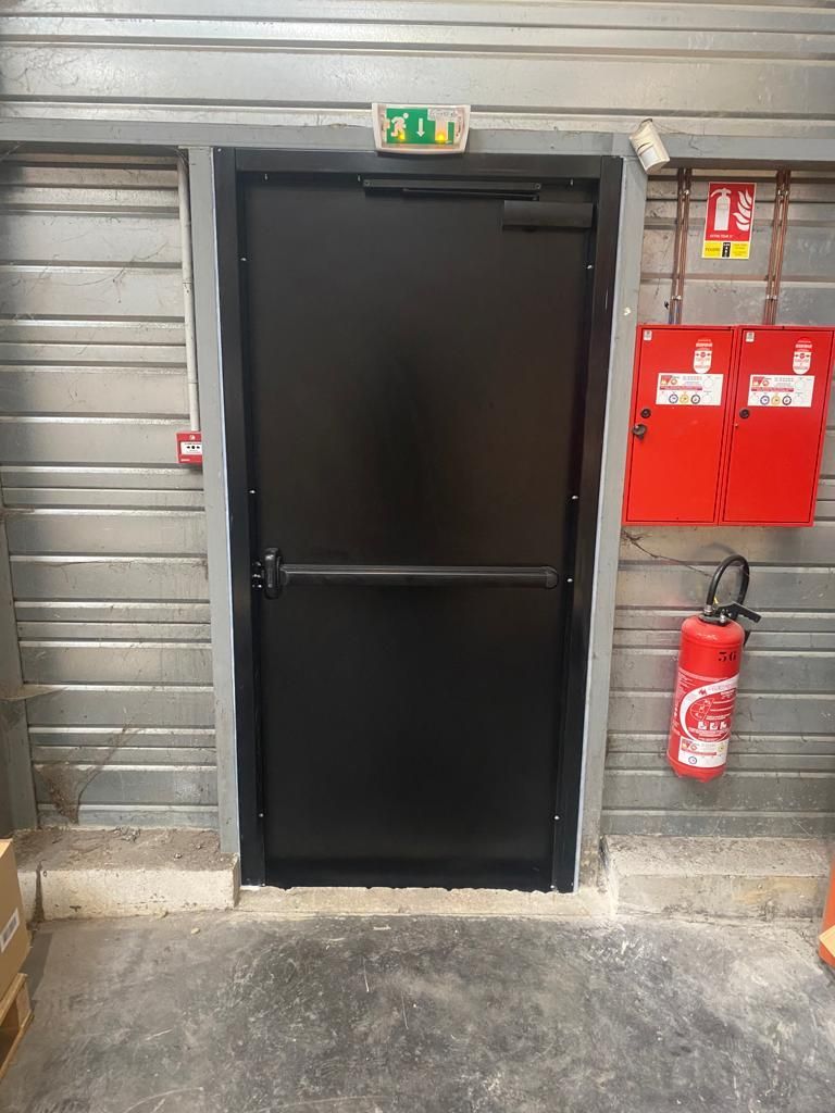 Porte en métal noir dans un mur industriel gris, avec des équipements et des panneaux de sécurité incendie à côté.