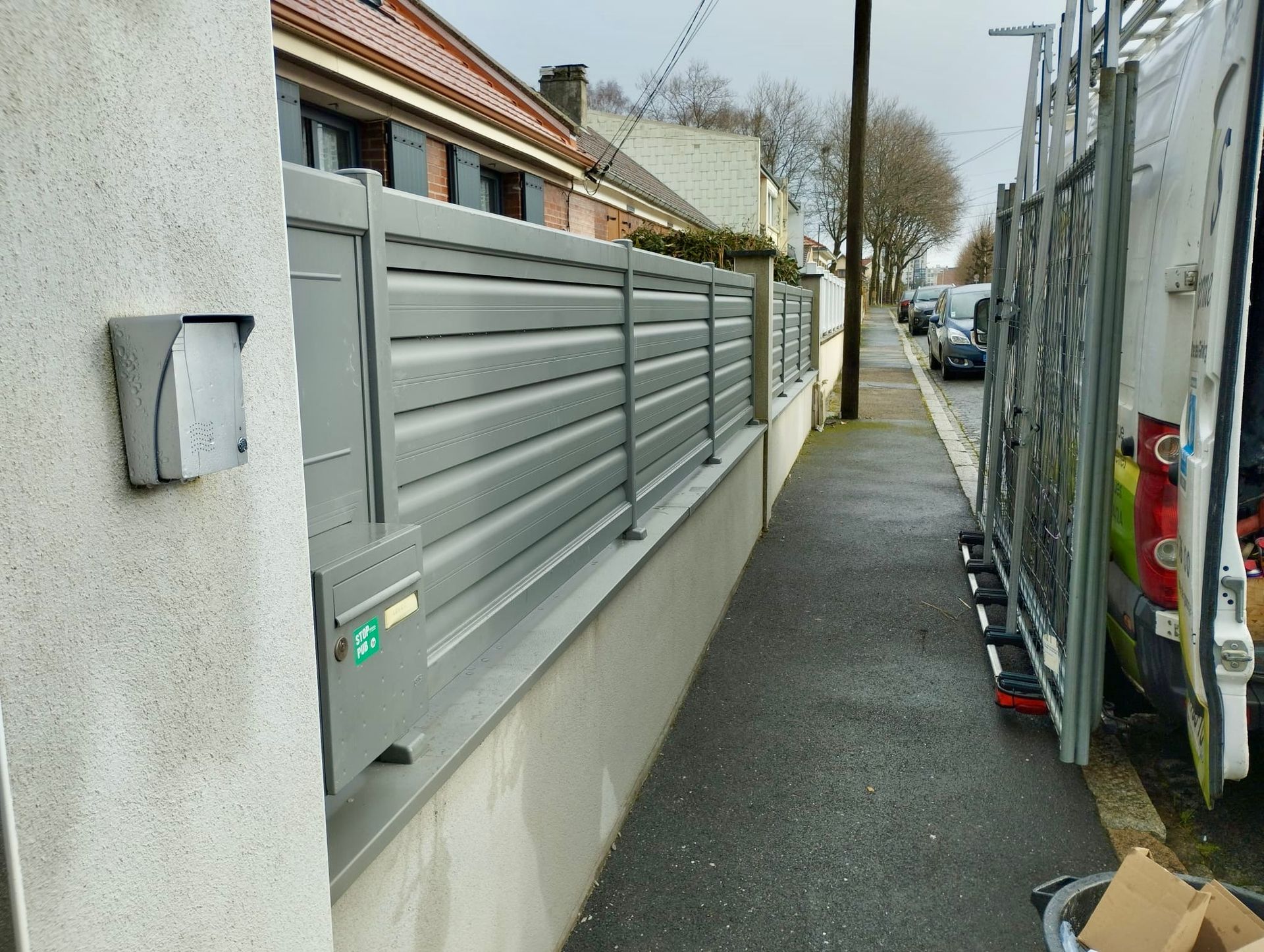 Ruelle étroite longeant une clôture grise et un bâtiment, avec une structure métallique et des véhicules stationnés sur la droite.