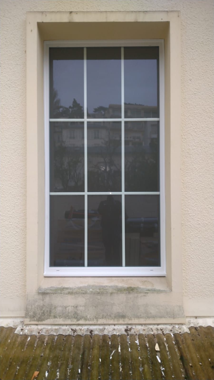 Haute fenêtre murale beige avec encadrement blanc et verre réfléchissant, au-dessus d'un rebord moussu.