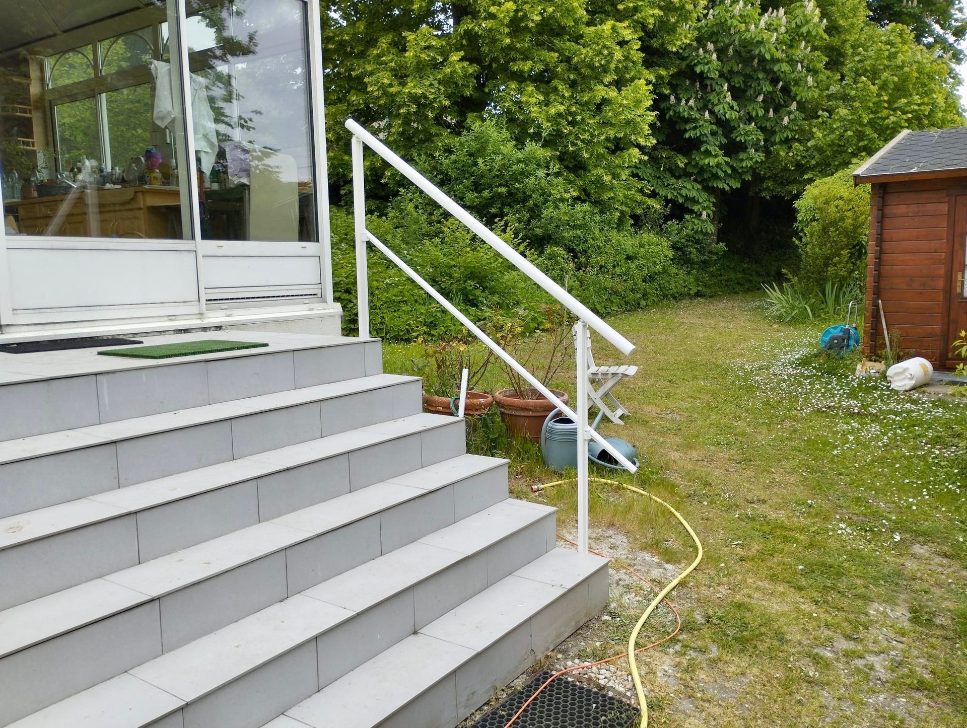 Un escalier blanc avec une rampe mène au porche d'une maison donnant sur une cour gazonnée et un petit abri de jardin.