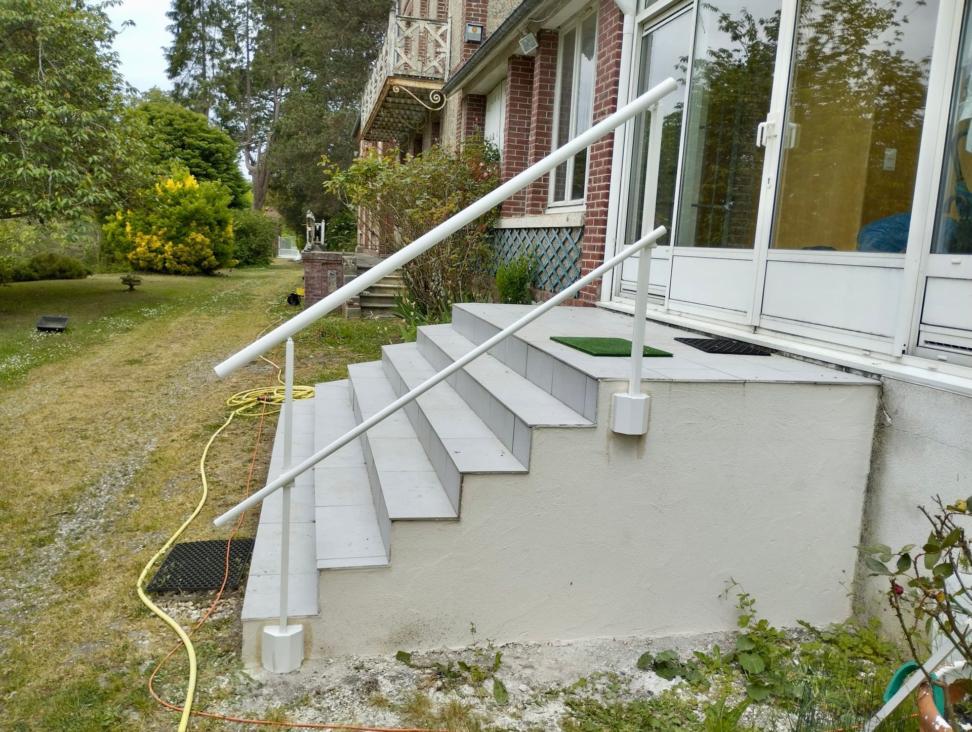 Un escalier avec une rampe blanche mène aux portes vitrées d'une maison en briques, à côté d'une cour gazonnée avec un tuyau d'arrosage jaune.