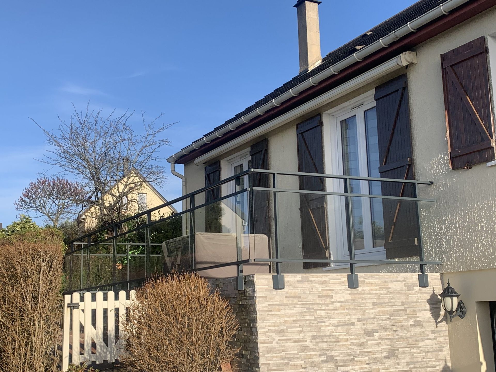 Extérieur de maison avec balustrade blanche, façade en pierre beige et volets sombres sous un ciel bleu