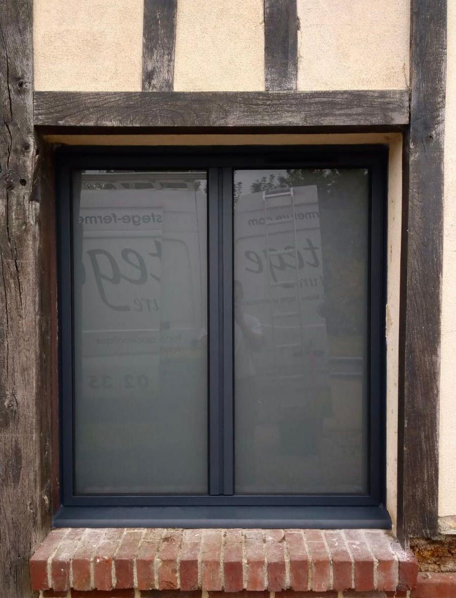 Une fenêtre à cadre sombre encastrée dans un mur beige, avec un appui en briques et du verre dépoli.
