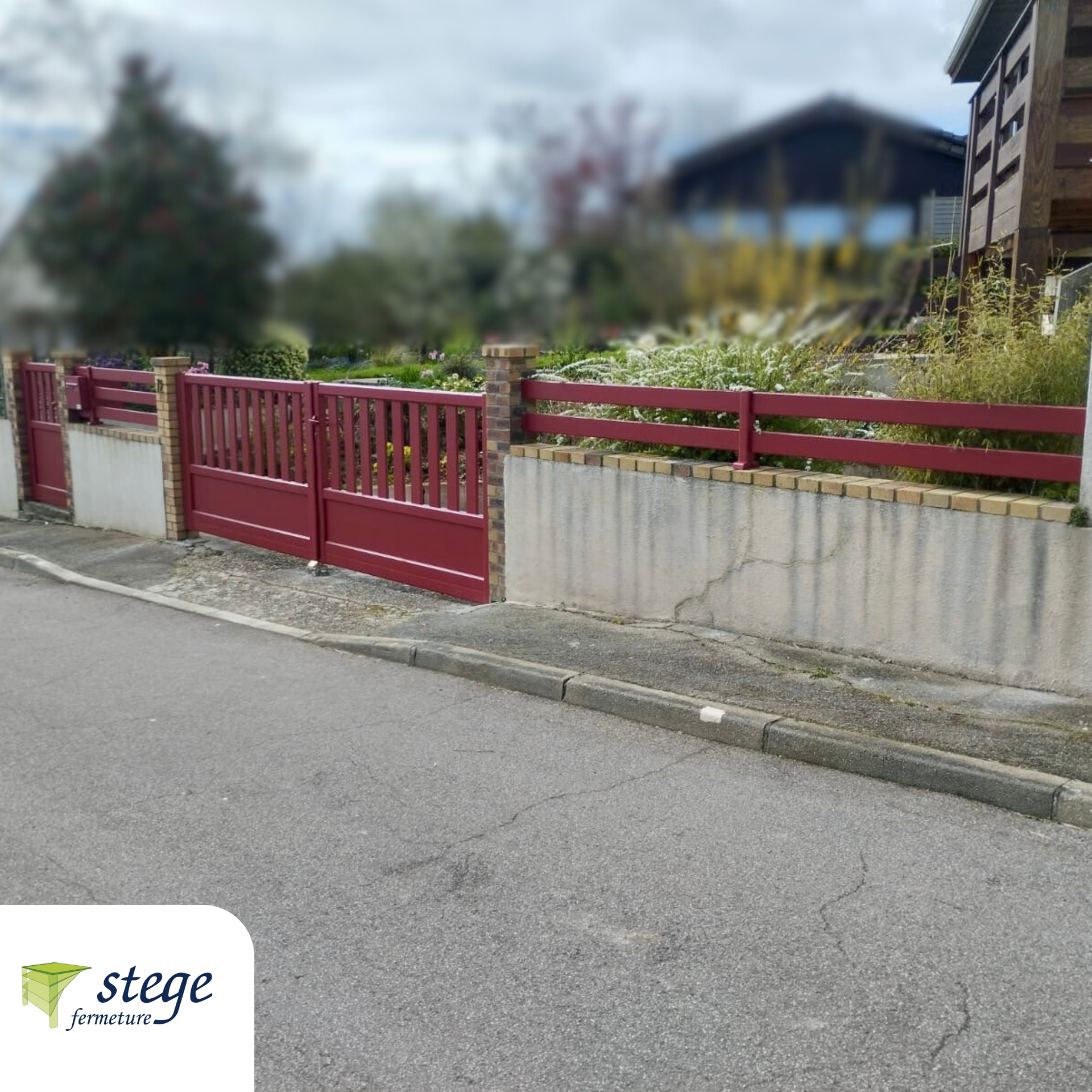Portail et clôture en métal rouge le long d'une rue tranquille, à côté d'un mur en béton et de buissons.