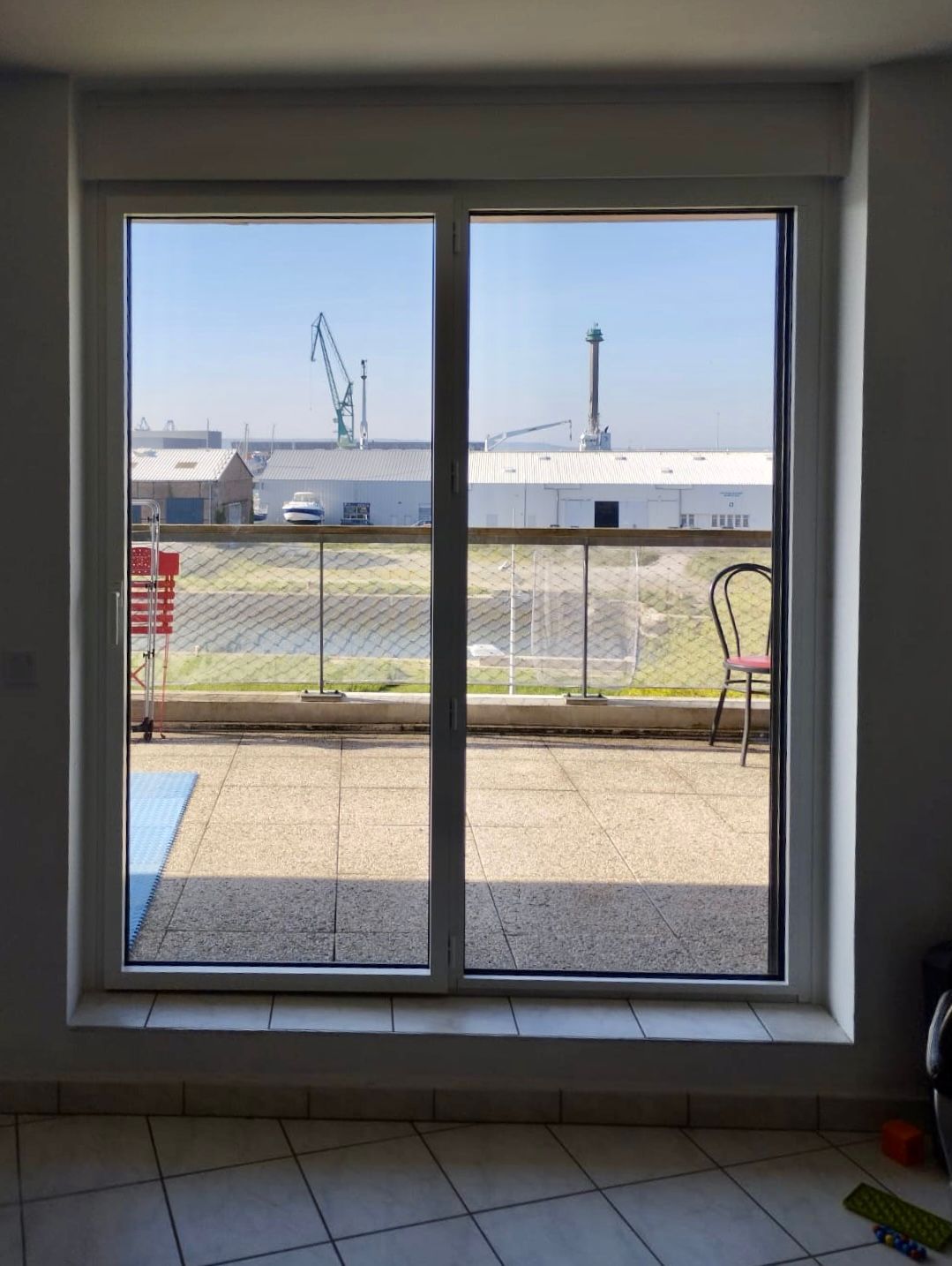Vue par une porte-fenêtre coulissante sur un patio ensoleillé, une clôture et des bâtiments industriels au-delà d'une zone herbeuse.