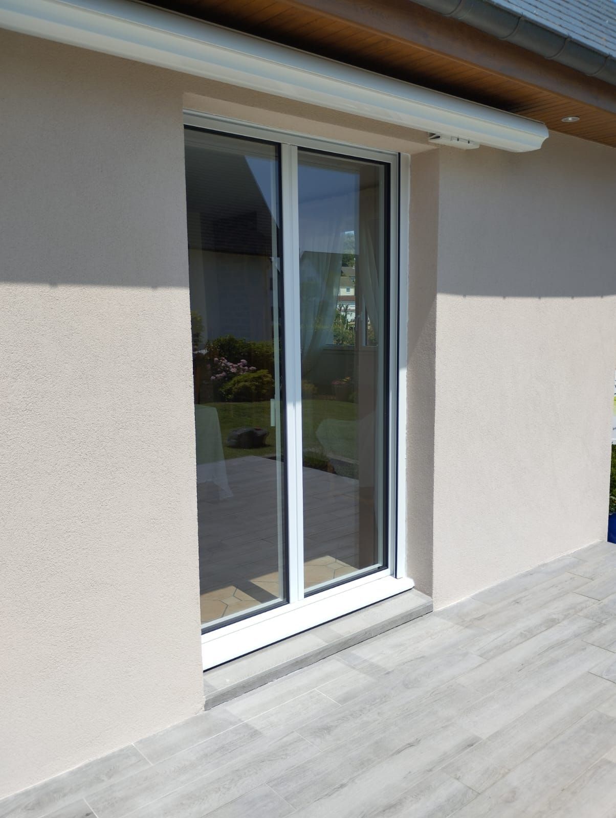Porte coulissante en verre beige sur un mur extérieur, avec un petit auvent au-dessus et une terrasse carrelée en dessous.