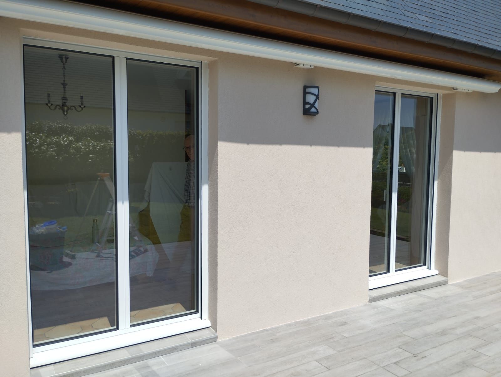 Mur extérieur avec deux portes coulissantes en verre et une applique murale donnant sur une terrasse beige.