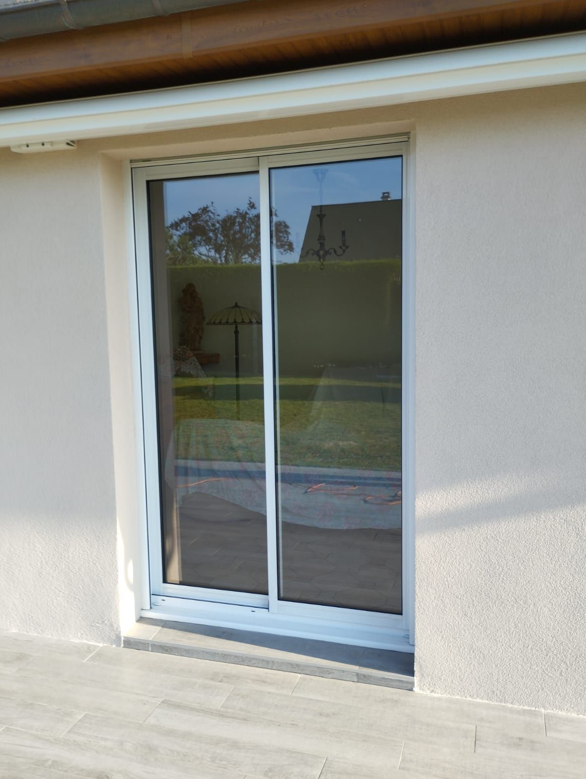 Porte coulissante en verre dans un mur en stuc blanc avec reflet sur le patio.