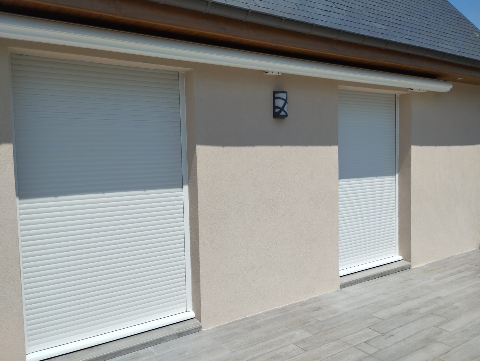 Deux volets blancs fermés sur un mur en stuc beige, à côté d'une terrasse pavée sous un toit de tuiles