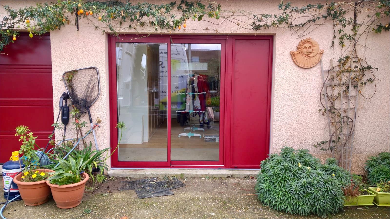 Entrée avec porte rouge et panneaux de verre, plantes en pot et lierre sur un mur en stuc.