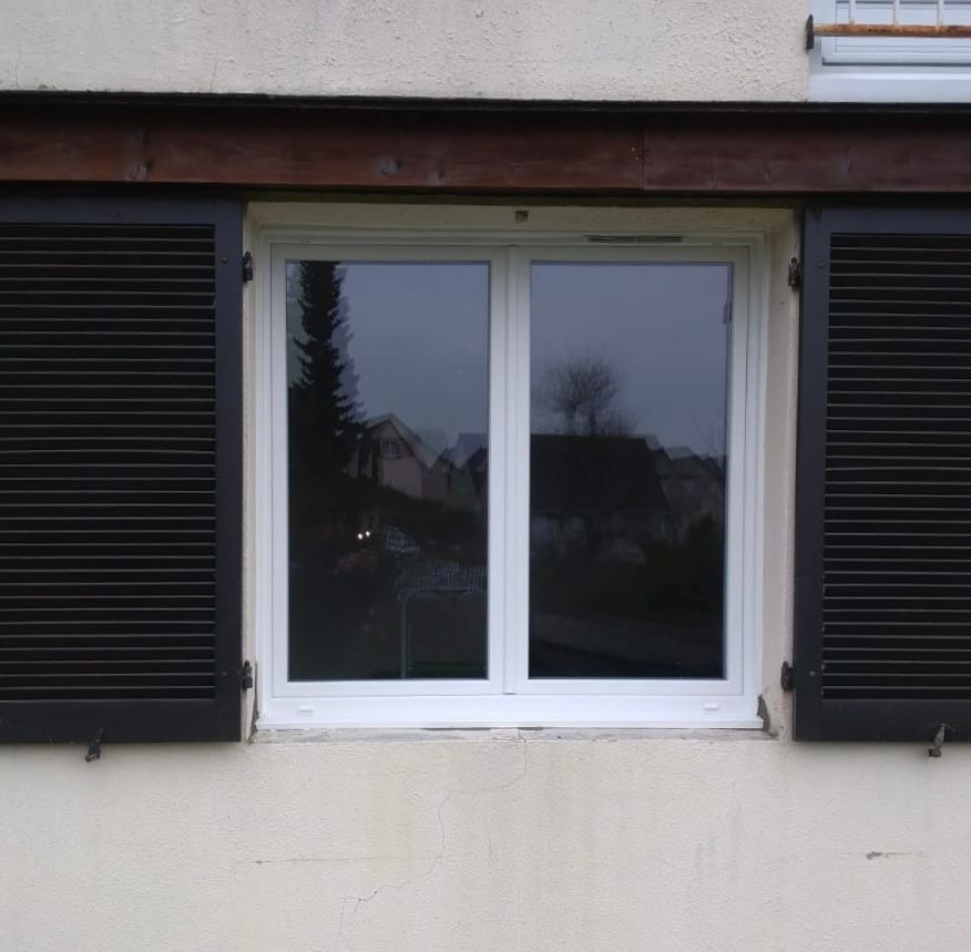 Fenêtre blanche à volets noirs sur un mur beige, reflétant des arbres et des maisons