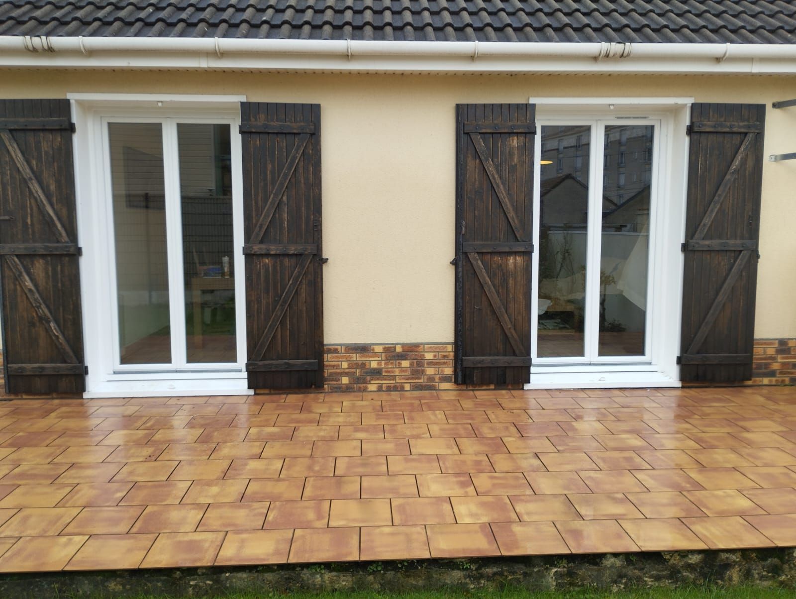 Extérieur de la maison avec fenêtres à cadre blanc, volets foncés et patio carrelé de terre cuite humide