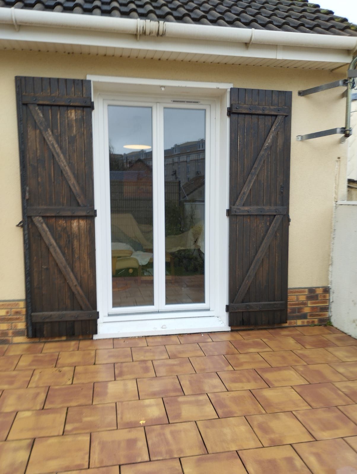 Portes-fenêtres blanches avec volets en bois foncé donnant sur une terrasse carrelée à l'extérieur d'une maison