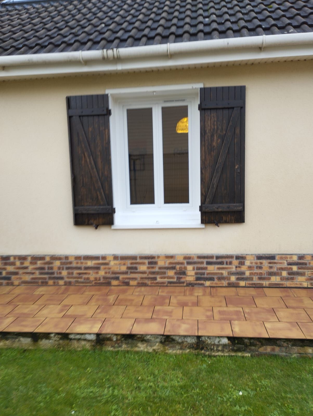 Extérieur de la maison avec fenêtre à cadre blanc, volets foncés et terrasse en briques donnant sur une pelouse.