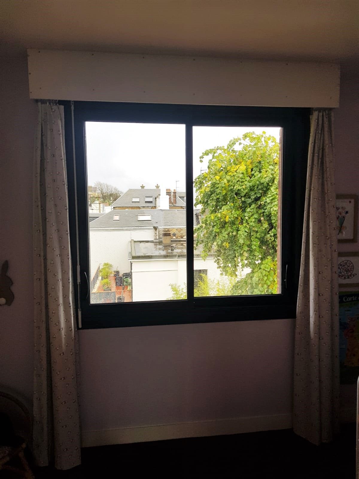 Fenêtre de chambre avec rideaux, donnant sur un arbre et des toits à l'extérieur.
