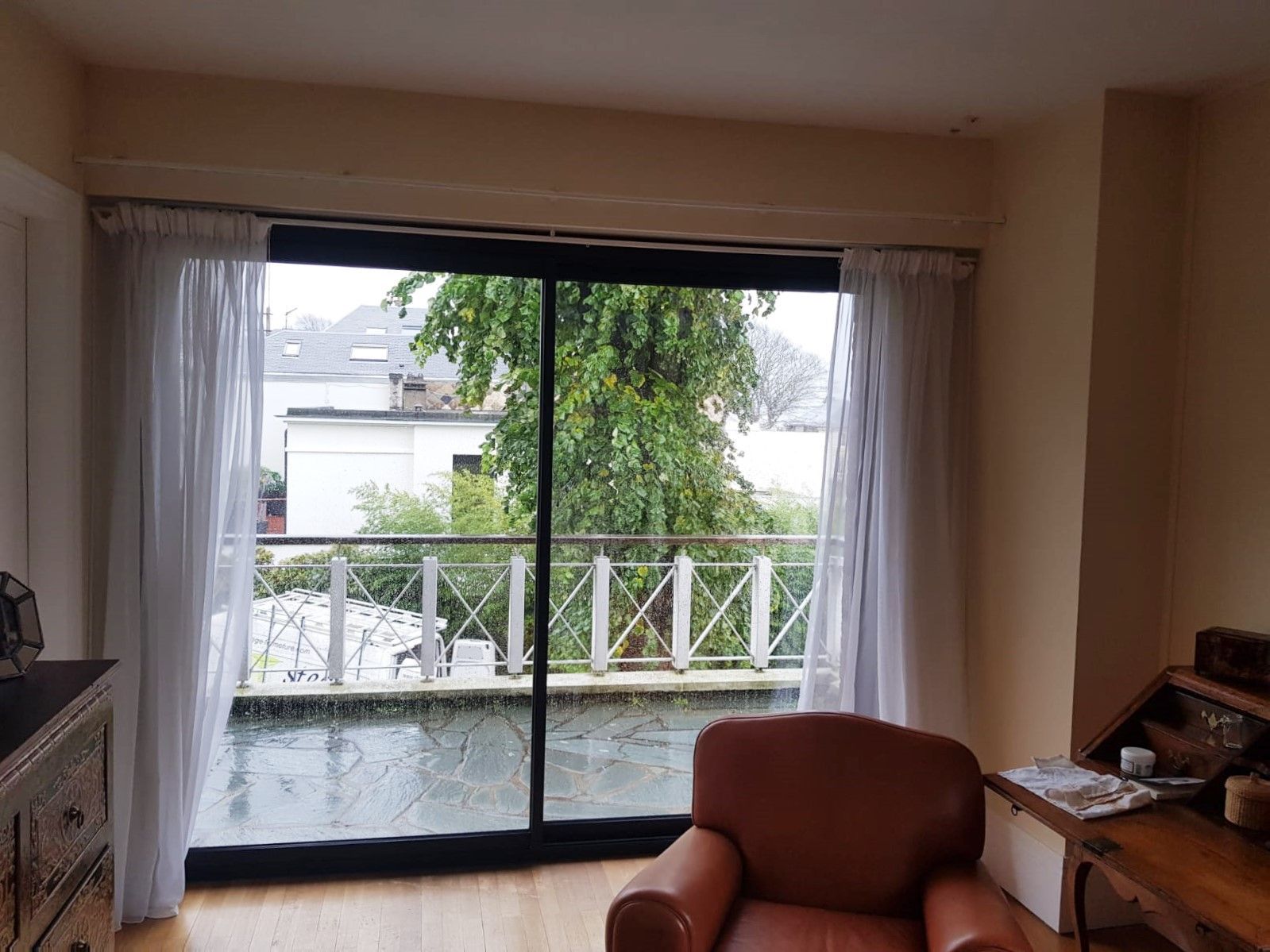 Salon avec portes coulissantes en verre, rideaux blancs et balcon donnant sur les arbres et les maisons
