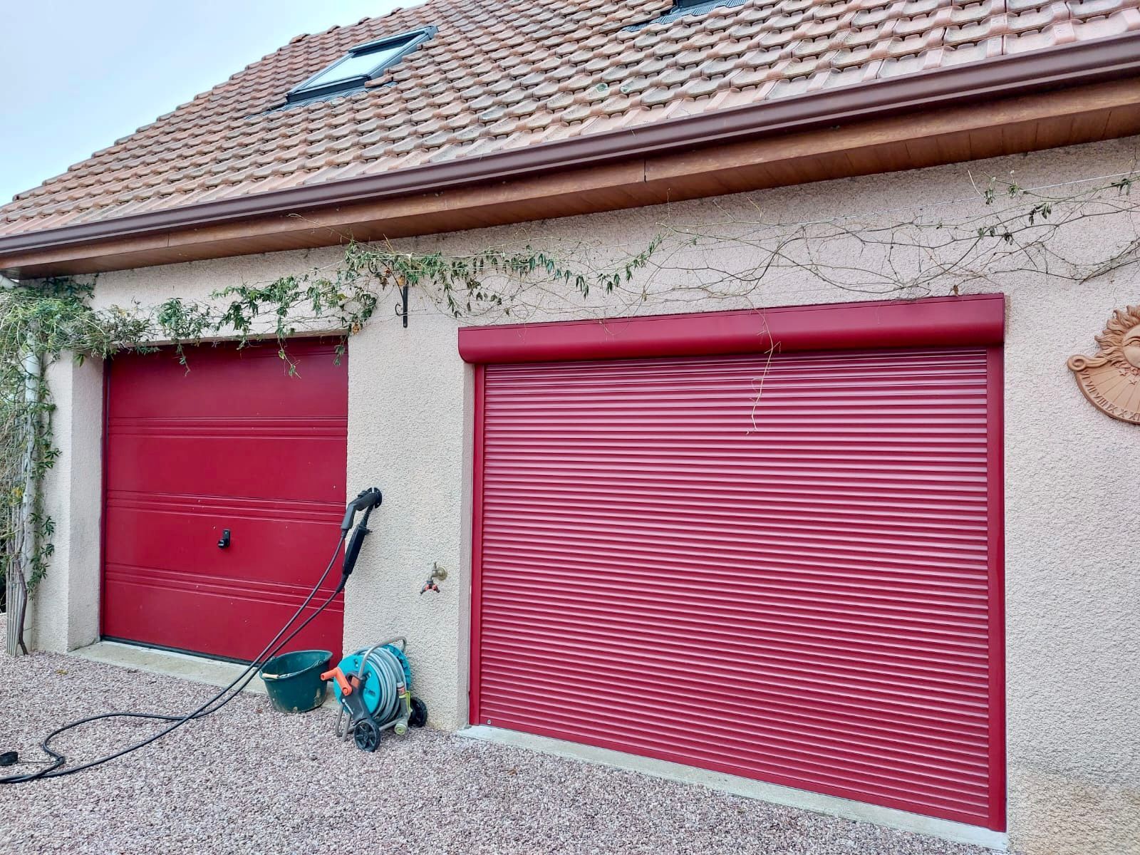 Portes de garage rouges sur une maison en stuc, avec un tuyau d'arrosage et du lierre le long du mur