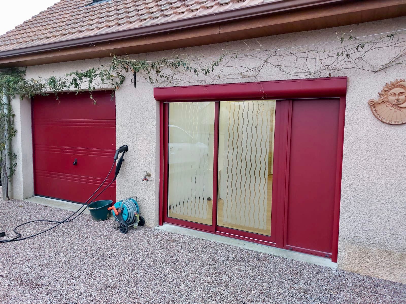 Extérieur d'une maison avec porte de garage rouge et porte-fenêtre coulissante donnant sur une allée en gravier