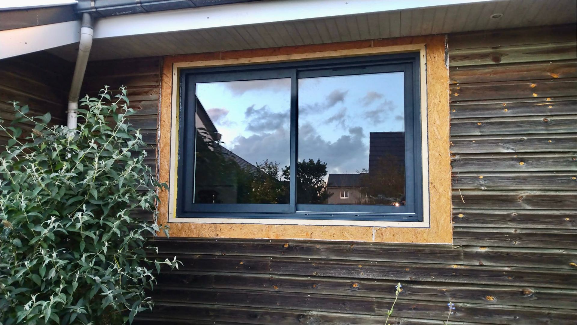 Façade en bois d'une maison avec une fenêtre à cadre sombre reflétant le ciel et les arbres environnants.