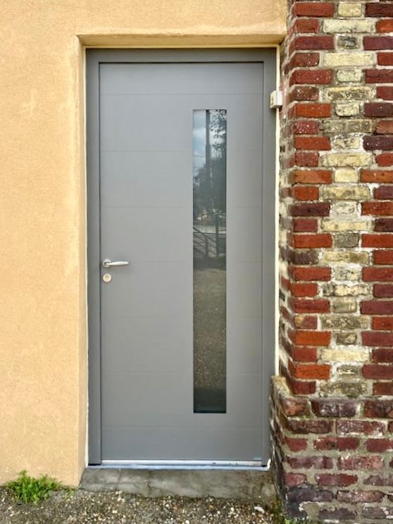Porte grise avec une étroite fenêtre verticale encastrée dans un mur de briques et de stuc