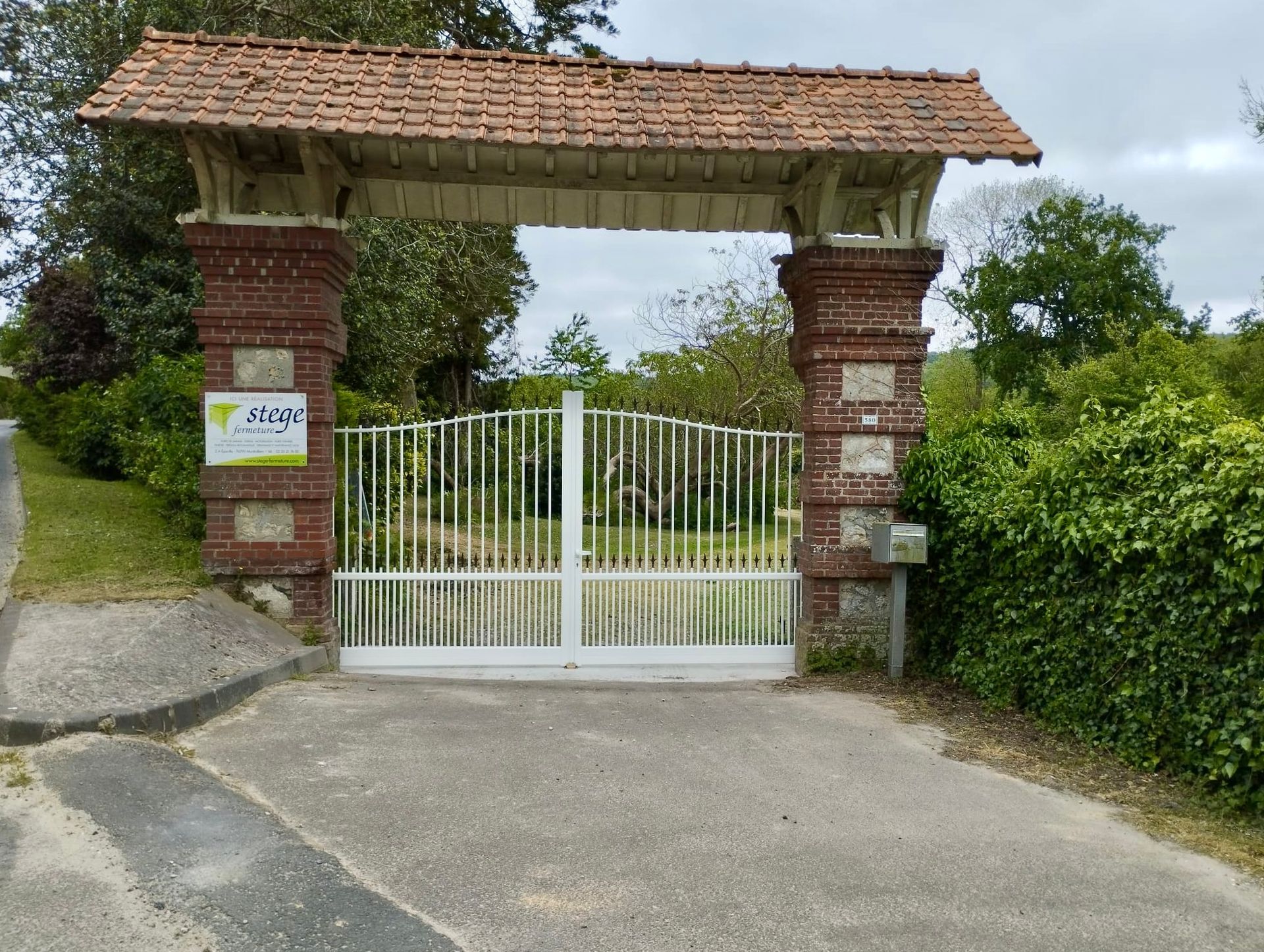 Portail blanc entre des piliers de briques avec un toit de tuiles à l'entrée d'une allée rurale