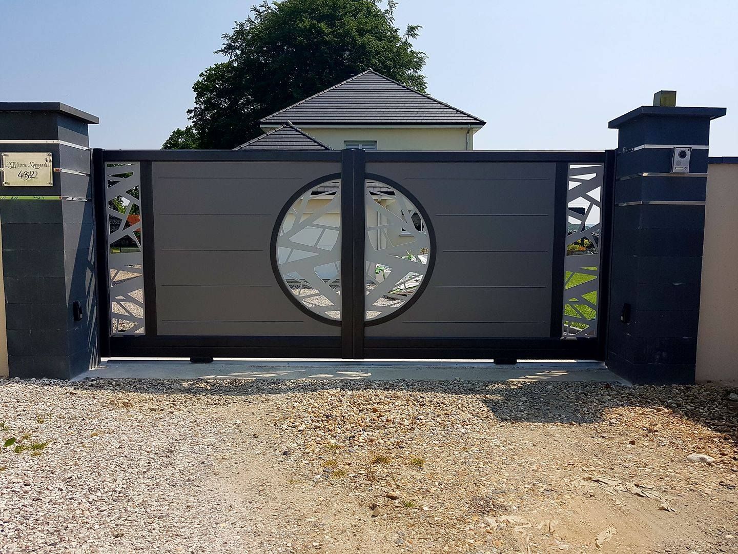 Portail moderne en métal noir avec un motif central circulaire, installé entre des piliers en pierre sur une allée de gravier.