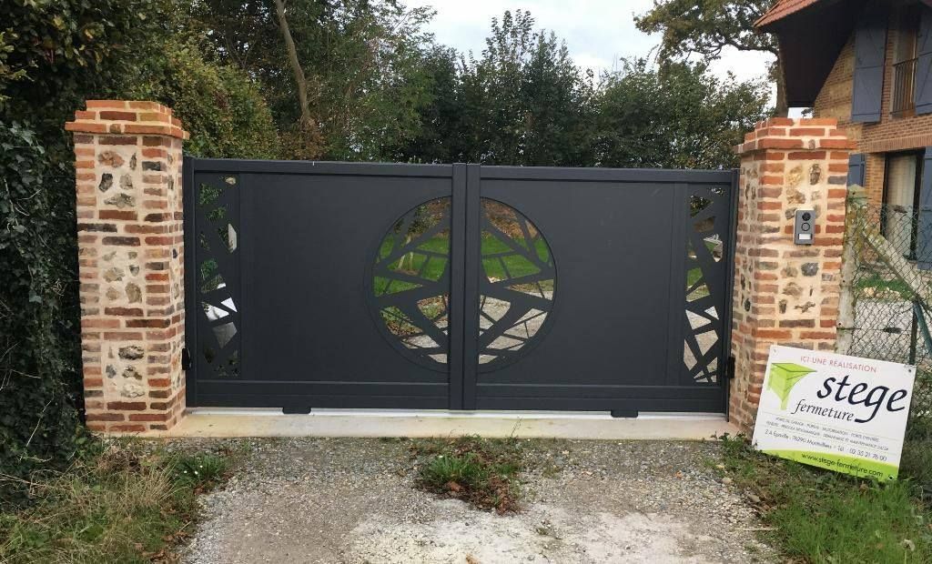 Portail d'allée en métal noir entre des piliers en briques, orné d'un motif d'arbre circulaire et d'un panneau de jardin sur la droite.
