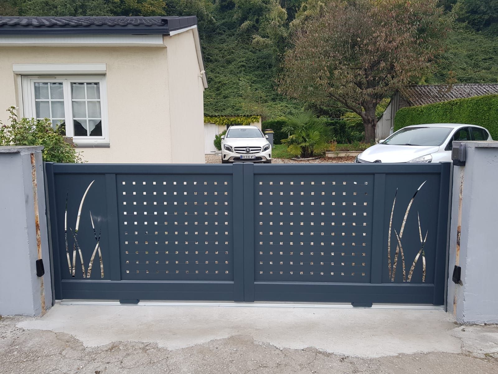 Portail d'allée en métal gris devant une maison avec deux voitures garées derrière.