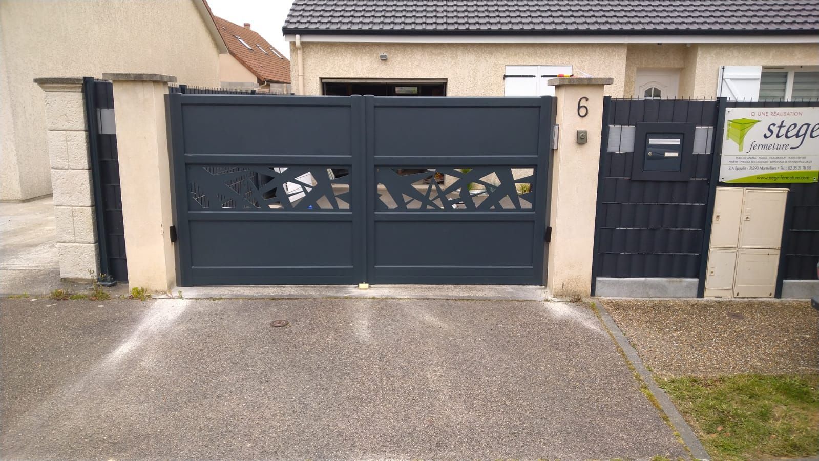 Portail d'allée gris foncé et porte piétonne à côté d'une maison beige et d'un panneau mural.
