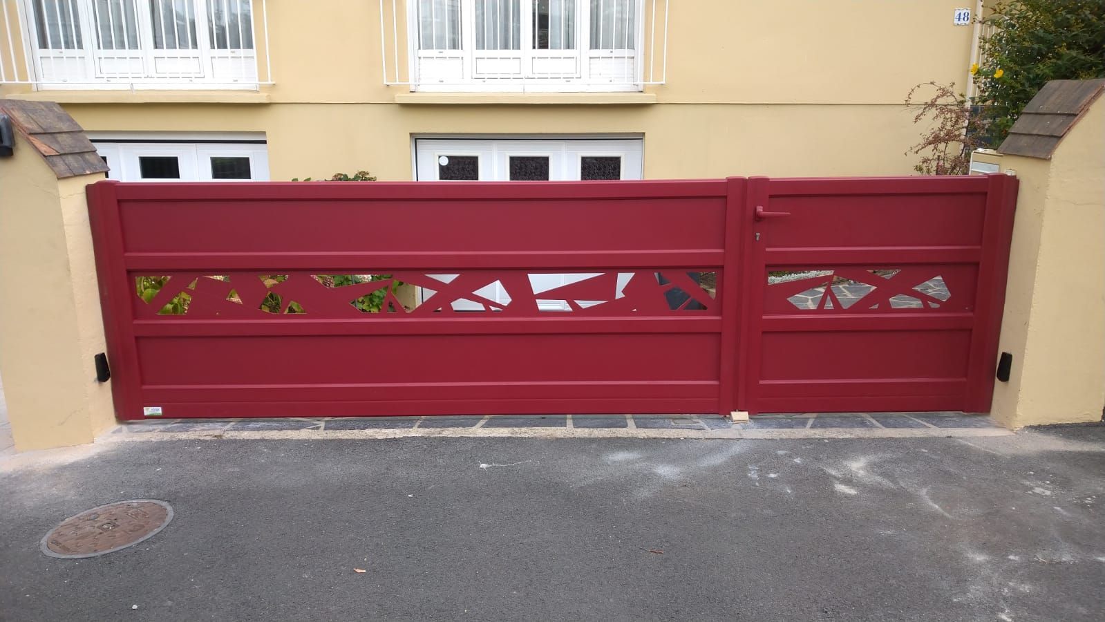 Portail d'allée en métal rouge devant un bâtiment beige avec fenêtres et balcons.