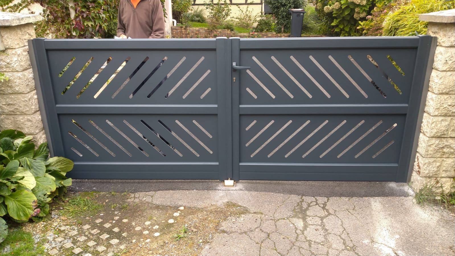 Portail d'allée en métal gris foncé avec des lattes diagonales et un loquet central, placé devant un chemin de pierre et un jardin.