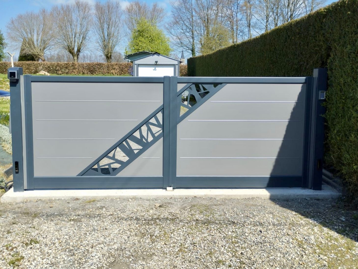 Portail coulissant moderne en métal gris avec motifs en treillis diagonal, installé devant des haies et des arbres