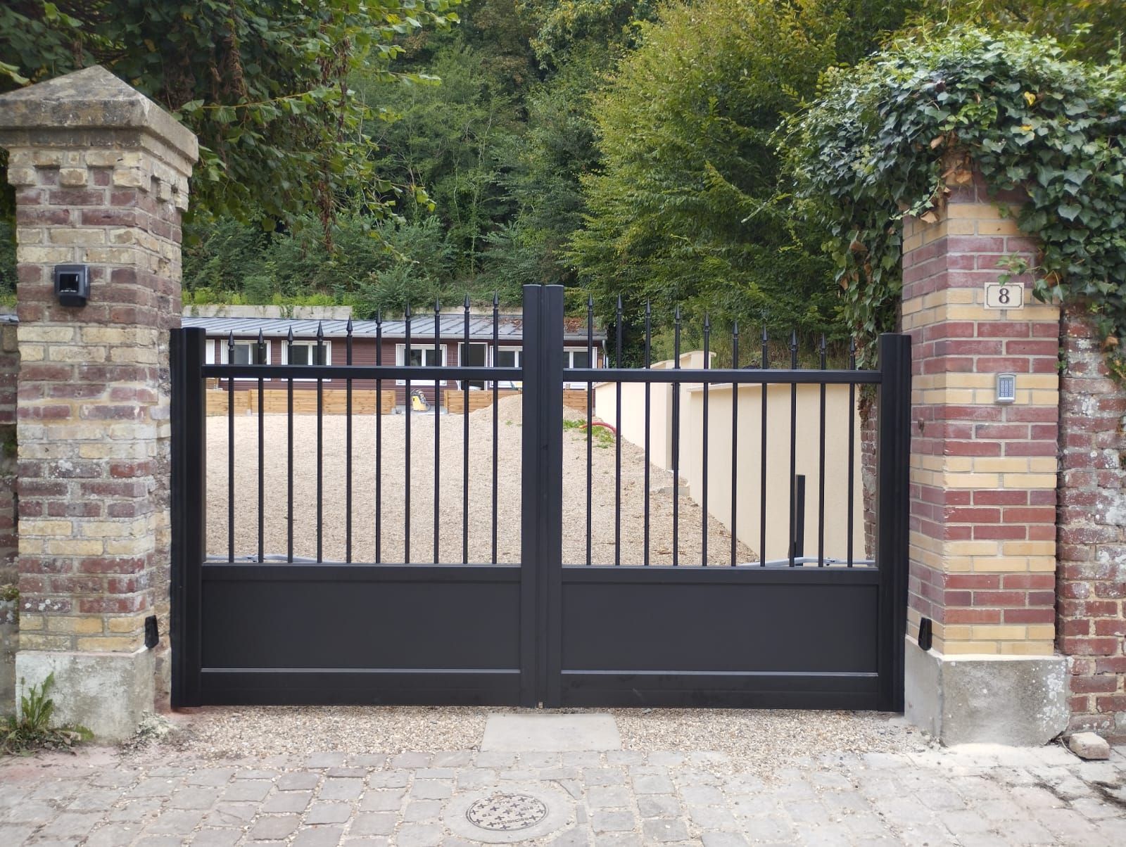 Portail d'allée en métal noir entre des piliers en briques à l'entrée d'un jardin